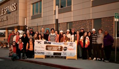 Many Parts, One Body volunteers and students gathered outside Blue Springs High School holding the organization's sponsor banner
